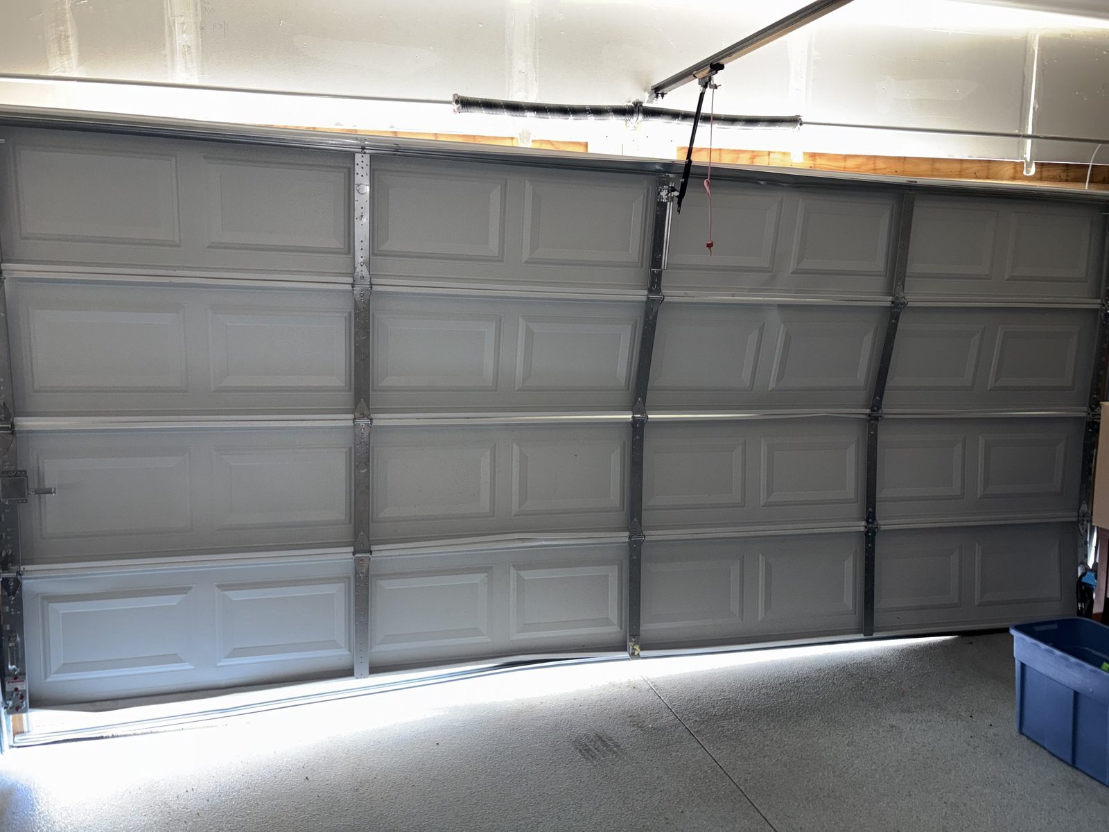 Inside a Colorado Springs garage: a white steel sectional door seen from behind, with its hinges, reinforcing struts, opener arm and the torsion spring above the header.