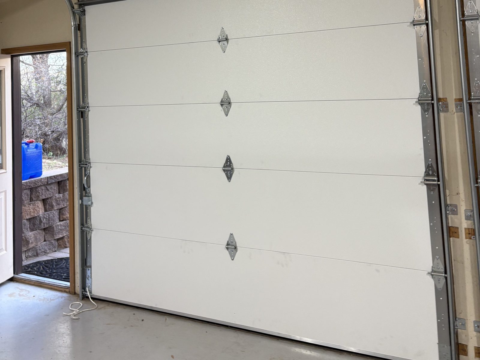 Inside a Colorado Springs garage in daylight: a new white sectional door closed to the floor, with the side door open to the yard.